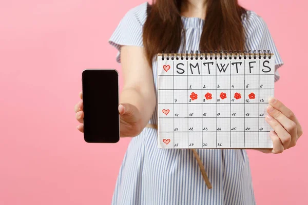 Woman holding female periods calendar for checking menstruation days, mobile phone with blank black empty screen isolated on pink background. Medical, healthcare, gynecological concept. Copy space