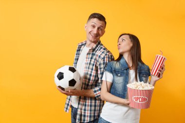 Genç eğlenceli çift, kadın erkek, holding futbol topu, kova patlamış mısır, plastik bardak kola, futbol taraftarları destek ekibi, sarı zemin üzerine izole neşelen. Spor aile eğlence yaşam tarzı kavramı