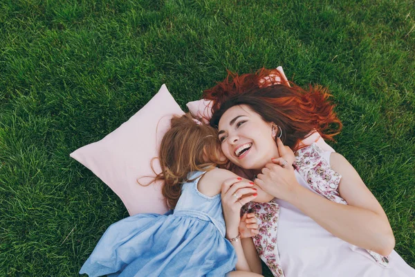 Laughing woman and little cute child baby girl lie on pillows on grass in park hug, embrace and rest, have fun. Mother, little kid daughter. Mother\'s Day, love family, parenthood, childhood concept