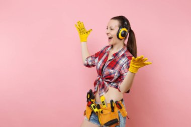 Tamirci kadını ekose gömlek, kot şort, sarı eldivenler, Ses Yalıtımlı kulaklık seti araçları, pembe arka plan izole çeşitli araçların tam kemer. Kadın erkek iş. Yenileme kavramı