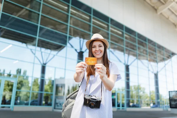 Young smiling traveler tourist woman in hat with backpack, retro vintage photo camera, holding credit card at international airport. Passenger traveling abroad on weekends getaway. Air flight concept