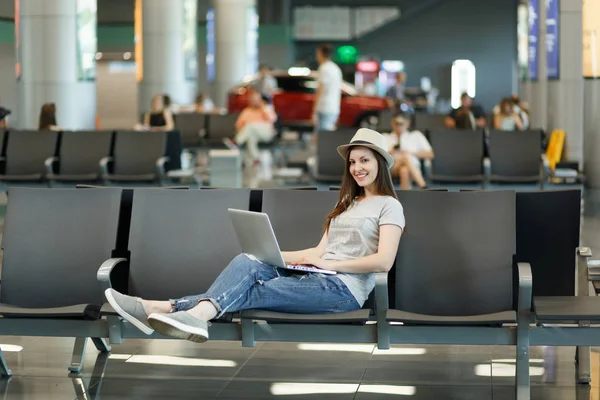Young smiling traveler tourist woman in hat working on laptop while waiting in lobby hall at international airport. Passenger traveling abroad on weekends getaway. Air travel, flight journey concept