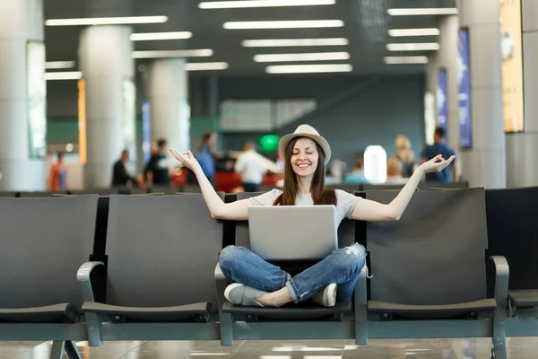 Young relaxed traveler tourist woman with laptop sitting with crossed legs, meditate, spread hands, waiting in lobby hall at airport. Passenger traveling abroad on weekend getaway. Air flight concept