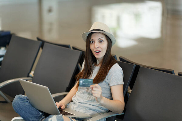 Молодая шокированная туристка в шляпе сидит, работает на ноутбуке, держит кредитку, ожидая в холле аэропорта. Пассажир, путешествующий за границу по выходным. Концепция полета
