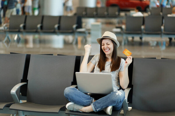 Радостный путешественник туристическая женщина с ноутбуком сидя со скрещенными ногами делать жест победителя держать кредитную карту ждать в вестибюле в аэропорту. Пассажир, выезжающий за границу в выходные. Концепция полета
