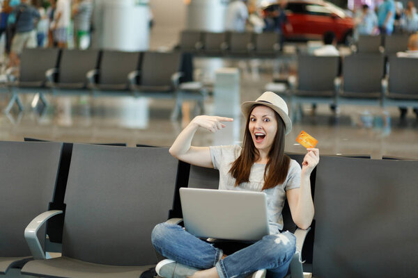 Потрясенная туристка в шляпе сидит со скрещенными ногами с ноутбуком, указывающим пальцем на кредитную карту, и ждет в холле аэропорта. Пассажир, выезжающий за границу в выходные. Концепция полета
