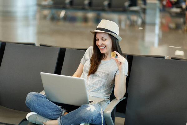 Молодая улыбчивая туристка в шляпе сидит со скрещенными ногами, работая на ноутбуке, держа биткойн в холле аэропорта. Пассажир, путешествующий за границу по выходным. Концепция полета
