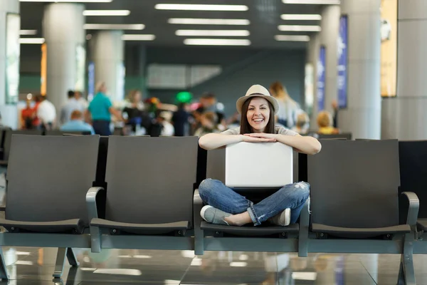 Young laughing traveler tourist woman sitting with laptop with crossed legs while waiting in lobby hall at international airport. Passenger traveling abroad on weekends getaway. Air flight concept