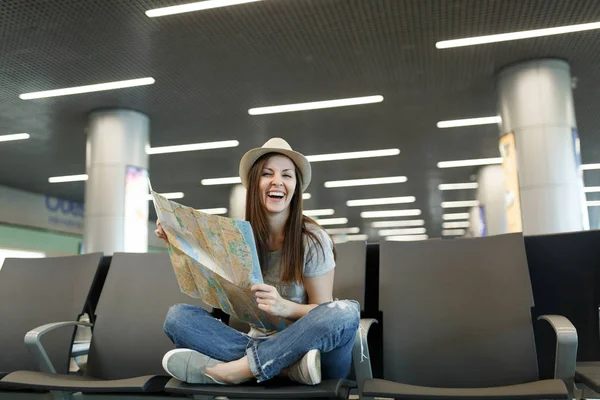 Young laughing traveler tourist woman with crossed legs hold paper map, search route waiting in lobby hall at international airport. Passenger traveling abroad on weekend getaway. Air flight concept