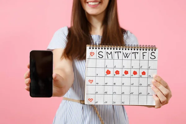 Woman holding female periods calendar for checking menstruation days, mobile phone with blank black empty screen isolated on pink background. Medical, healthcare, gynecological concept. Copy space