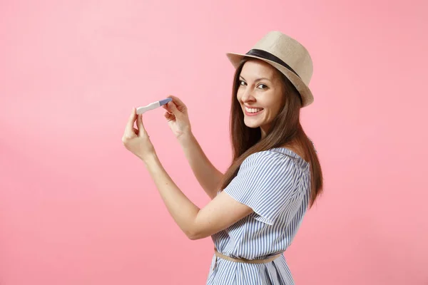 Excited happy woman in blue dress, hat hold in hand, looking at pregnancy test isolated on pink background. Medical healthcare gynecological, pregnancy fertility maternity people concept. Copy space