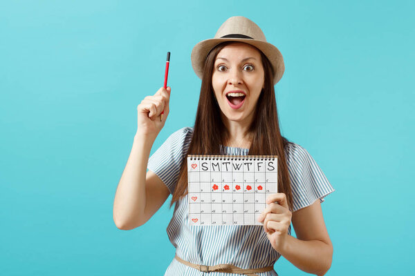 Portrait of happy woman in blue dress, hat holding red pencil, female periods calendar for checking menstruation days isolated on blue background. Medical healthcare gynecological concept. Copy space