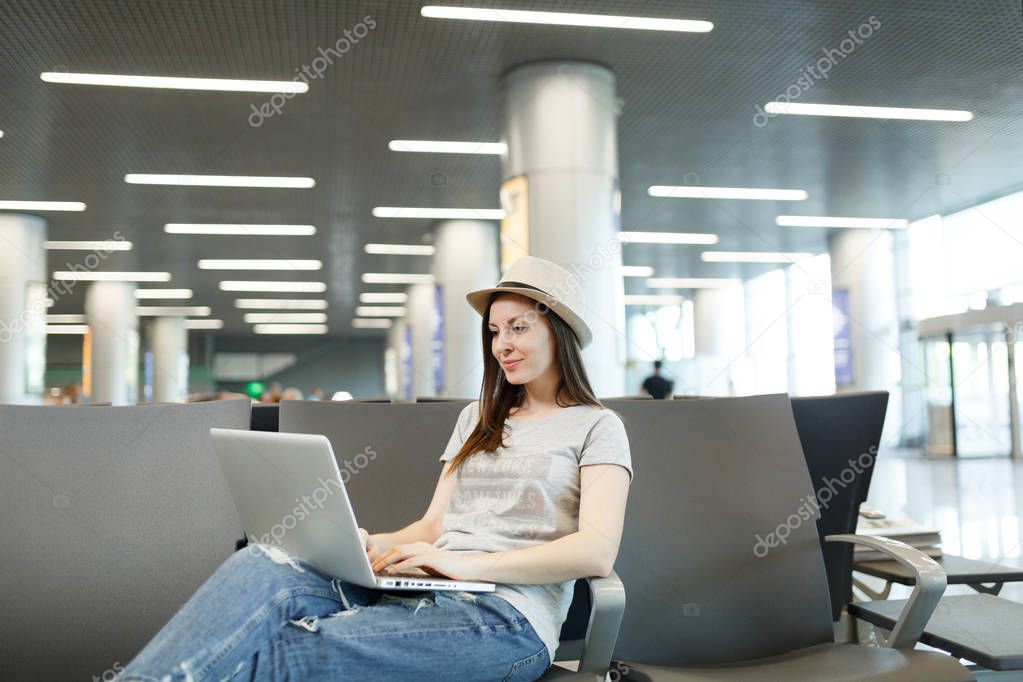 Young calm traveler tourist woman in hat sitting, working on laptop, waiting in lobby hall at international airport. Passenger traveling abroad on weekend getaway. Air travel, flight journey concept