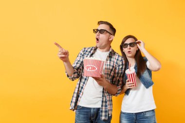 Genç çift kadın adam 3d gözlük kova patlamış mısır plastik bardak kola veya kola işaret parmağı bir kenara izole sarı arka plan üzerinde tutarak tarihi film film izlerken şok. Sinema duygular