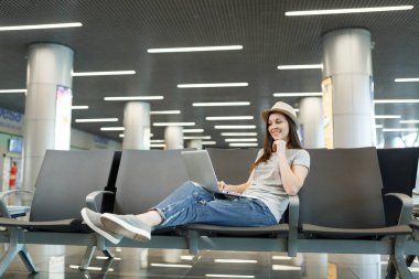 Şapkalı lobi salonunda uluslararası havaalanında beklerken dizüstü bilgisayar üzerinde çalışan genç sevimli seyahat turizm kadın. Hafta sonu kaçamağı yurt dışında seyahat yolcu. Hava seyahat, uçuş yolculuk kavramı