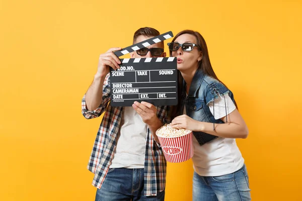 Young happy funny couple woman man in 3d glasses watching movie film on date holding classic black film making clapperboard bucket of popcorn isolated on yellow background. Emotions in cinema concept