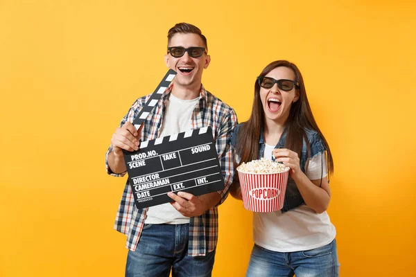 Young joyful couple woman man in 3d glasses watching movie film on date holding classic black film making clapperboard and bucket of popcorn isolated on yellow background. Emotions in cinema concept