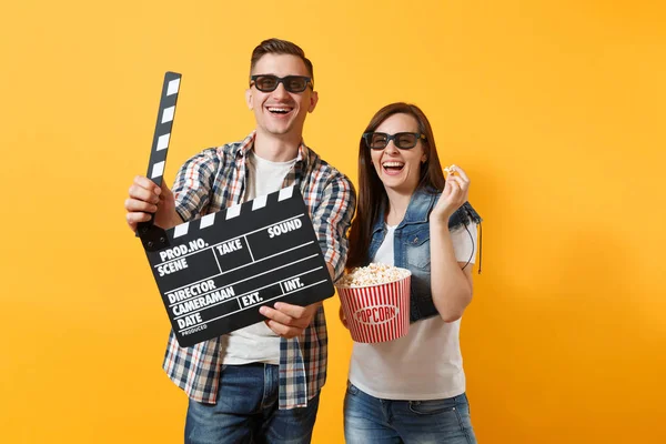 Young laughing couple woman man in 3d glasses watching movie film on date holding classic black film making clapperboard bucket of popcorn isolated on yellow background. Emotions in cinema concept