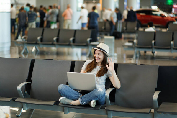 Путешественница-туристка в шляпе работает на ноутбуке, машет рукой для приветствия в веб-камере во время видеозвонка, ждет в холле аэропорта. Пассажир, путешествующий за границу по выходным. Концепция полета
