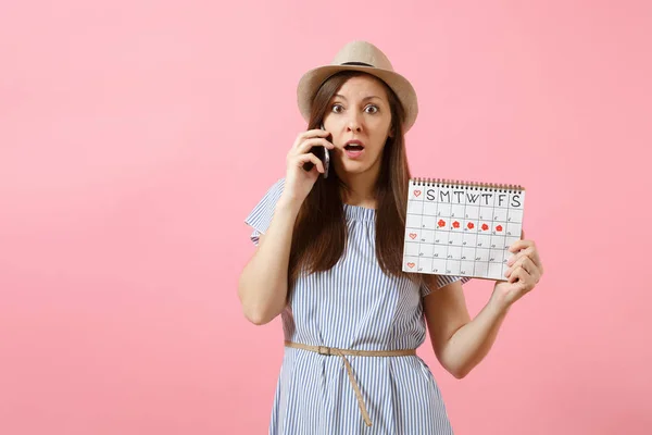 Sad confused woman talking on mobile phone, holding periods calendar for checking menstruation days isolated on bright trending pink background. Medical, healthcare, gynecological concept. Copy space