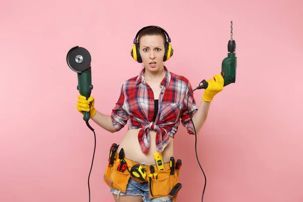 能源修理工妇女在手套 隔音耳机 套件工具带充满电力锯电动钻头在粉红色的背景上隔离 女性在男性工作 装修理念 — 图库照片