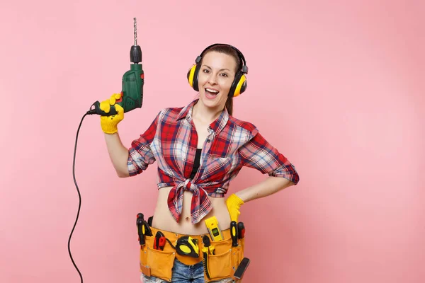Energy sexy handyman woman in gloves, noise insulated headphones, kit tools belt full of instruments holding power electric drill isolated on pink background. Female in male work. Renovation concept