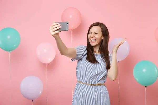Portrait of laughing pretty woman in blue dress doing selfie on mobile phone spreading hands on pink background with colorful air balloons. Birthday holiday party, people sincere emotions concept