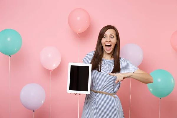 Portrait of excited young woman in blue dress holding pointing index finger on tablet pc computer with blank empty screen on pink background with colorful air balloons. Birthday holiday party concept