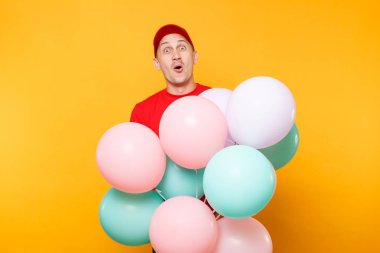 Kırmızı üniformalı teslimatçı sarı arka planda izole edilmiş. Profesyonel erkek çalışan şapkalı, tişörtlü kurye satıcısı elinde renkli pastel renkli hava balonları tutuyor. Hizmet kavramı. Boşluğu kopyala