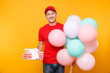 Delivery man in red uniform isolated on yellow orange background. Male employee in cap, t-shirt courier dealer holding bunch of colorful pastel air balloons, red gift box. Service concept. Copy space clipart