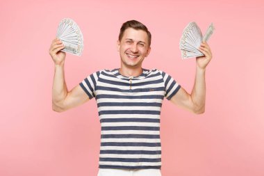 Gülümseyen portresi heyecanlı çizgili tişört holding pakette bulunan genç adam çok dolar, nakit para, şevk jest üstünde pembe arka plan izole kopya yer. İnsanlar samimi duygular yaşam tarzı kavramı