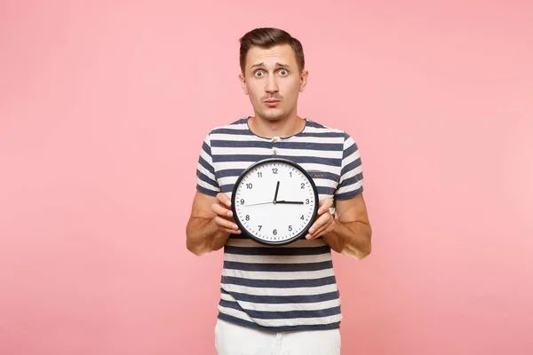 Portrait of shocked sad upset man wearing striped t-shirt holding round clock, copy space isolated on trending pastel pink background. People sincere emotions lifestyle concept. Time is running out