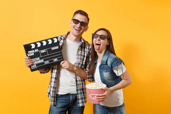 Young joyful couple woman man in 3d glasses watching movie film on date holding classic black film making clapperboard and bucket of popcorn isolated on yellow background. Emotions in cinema concept