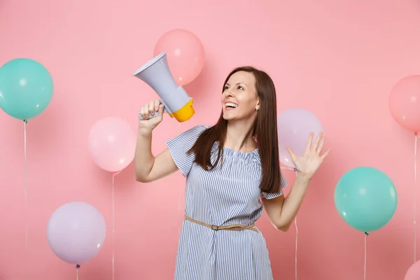 Portrait of joyful young attaractive woman wearing blue dress holding megaphone spreading hands on pink background with colorful air baloons. Birthday holiday party, people sincere emotions concept