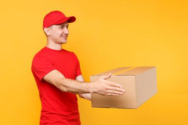 Teslimatçı sarı turuncu arka plan üzerinde izole kırmızı üniforma. Profesyonel erkek çalışan kurye veya Bayi kap t-shirt boş karton kutu tutarak. Paket hizmet anlayışı alma.