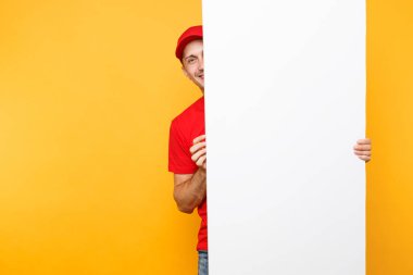 Teslimatçı sarı turuncu arka plan üzerinde izole kırmızı üniforma. Cap, t-shirt holding büyük beyaz boş boş billboard kopya alanı ile metin resim si erkek çalışan kurye. Hizmet anlayışı