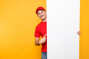 Teslimatçı sarı turuncu arka plan üzerinde izole kırmızı üniforma. Cap, t-shirt holding büyük beyaz boş boş billboard kopya alanı ile metin resim si erkek çalışan kurye. Hizmet anlayışı