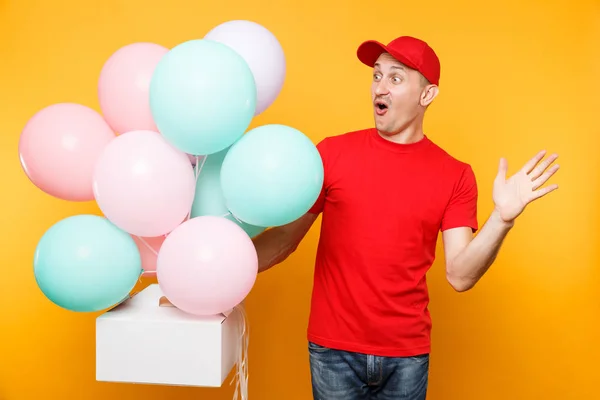 Man giving food order cake box isolated on yellow background. Male employee courier in red cap t-shirt hold colorful air balloons, dessert in empty cardboard box. Delivery service concept. Copy space