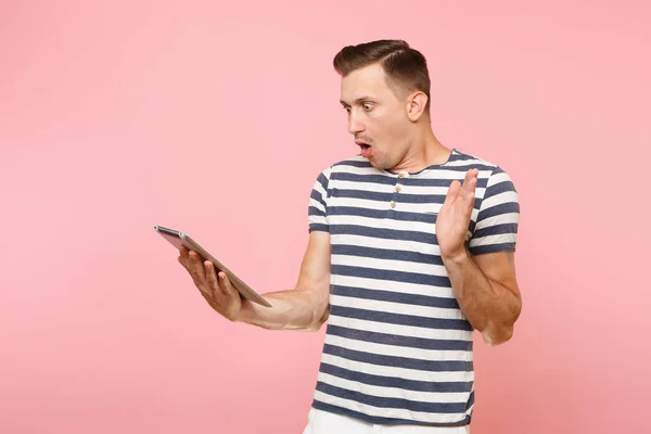 Portrait of shocked surprised user man in striped t-shirt working on tablet pc computer isolated on pastel pink background. People sincere emotions lifestyle concept. Advertising area on copy space