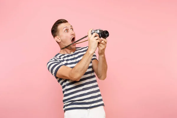 Portrait of surprised young photographer man wearing striped t-shirt take pictures on retro vintage photo camera isolated on trending pastel pink background. People sincere emotions lifestyle concept