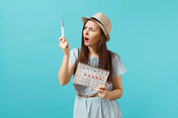 Shocked sad woman in blue dress, hat hold in hand pregnancy test, periods calendar for checking menstruation days isolated on blue background. Medical, healthcare, gynecological concept. Copy space