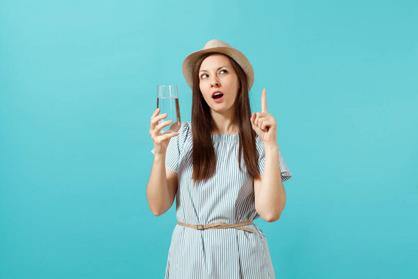 Portrait of young pensive woman in blue dress, hat holding and drinking clear fresh pure water from glass isolated on blue background. Healthy lifestyle, people sincere emotions concept. Copy space
