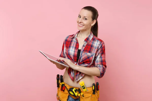 Young handyman woman in plaid shirt, denim shorts, kit tools belt full of variety instruments hold tablet pc computer money isolated on pink background. Female doing male work. Renovation concept