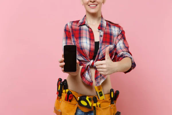 Woman with kit tools belt full of instruments holding showing camera mobile phone with blank black empty screen display touchscreen isolated on pink background. Female male work. Renovation concept