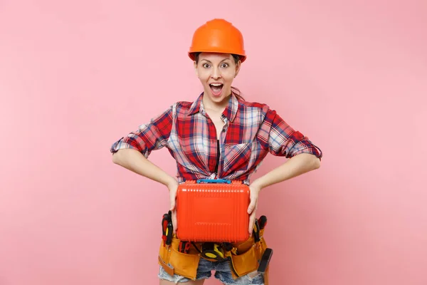 Strong young handyman woman in orange helmet, plaid shirt, denim shorts, kit tools belt full of instruments, toolbox isolated on pink background. Female in male work. Renovation occupation concept