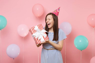 Doğum günü şapkası ve mavi elbise hediye ile renkli hava baloons pastel pembe arka plan üzerinde kırmızı kutu tutan çok memnun genç kadın portresi. Doğum günü tatil partisi, insanlar samimi duygular