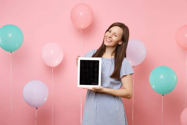 Portrait of happy smiling young woman in blue dress holding tablet pc computer with blank empty screen blinking on pastel pink background with colorful air balloons. Birthday holiday party concept
