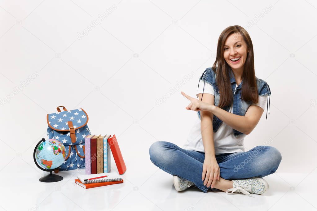 Portrait of young casual laughing woman student in denim clothes sitting pointing index finger on globe backpack school books isolated on white background. Education in high school university college