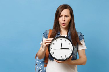 Sırt çantası ile denim giysili genç şaşkın kadın öğrenci portresi çalar saat izole mavi arka plan üzerinde tutun. Zaman tükeniyor. Üniversitesi'nde eğitim. Kopya alanı reklam için