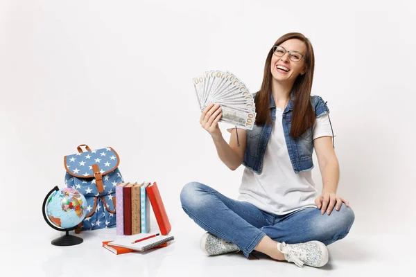年轻的笑女学生戴着眼镜捧着一捆大把的钞票 钱坐在地球仪上 书包里 学校里的书被隔离在白色的背景上 高中院校教育 — 图库照片